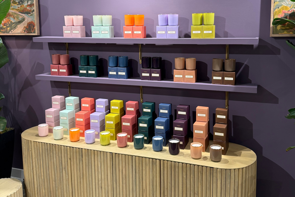 A retail display of many scented candles in colorful boxes and tins arranged on purple shelves and a light wood counter against a dark purple wall, with framed wall art and green plants partially visible.